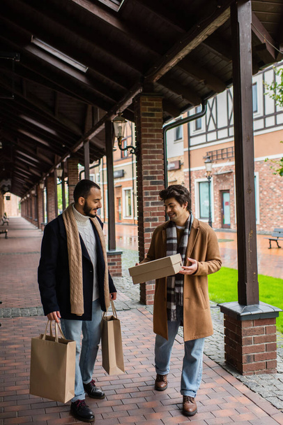 full length of happy gay man holding shoebox near bearded boyfriend with shopping bags on urban street - Photo, Image