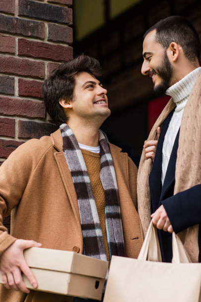 joyful and trendy gay couple with purchases smiling at each other near brick wall outdoors - Фото, зображення