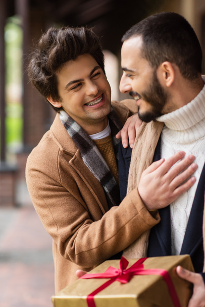 νέος και χαρούμενος ομοφυλόφιλος άνδρας αγγίζοντας το στήθος του γενειοφόρου φίλου κρατώντας χριστουγεννιάτικο δώρο σε εξωτερικούς χώρους - Φωτογραφία, εικόνα