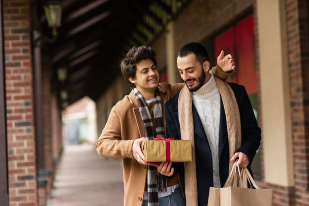 χαρούμενα γκέι άνδρας που παρουσιάζει κουτί δώρου στην ευχάριστη και μοντέρνα φίλο κρατώντας τσάντες ψώνια στην οδό της πόλης - Φωτογραφία, εικόνα