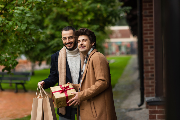 cheerful and fashionable gay men with christmas present and shopping bags looking at camera outdoors - Photo, Image