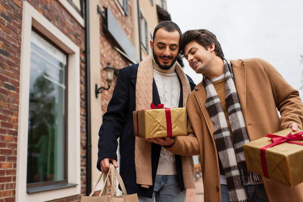 young gay man holding gift boxes near happy and trendy boyfriend with shopping bags on urban street - Фото, изображение