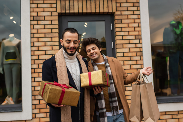 stylisches homosexuelles Paar hält Weihnachtsgeschenkboxen und Einkaufstüten in der Nähe des Geschäfts mit Vitrinen im Hintergrund - Foto, Bild