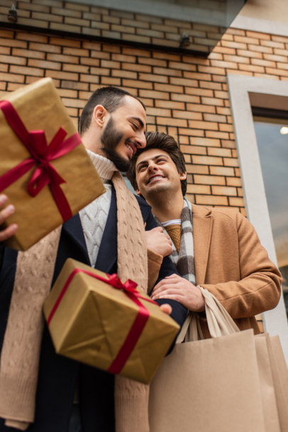 クリスマスプレゼント付きの明るいゲイ男性の低角度ビューや路上で建物の近くのショッピングバッグ - 写真・画像
