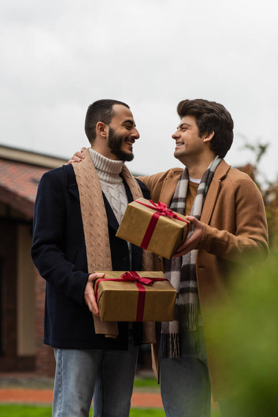 trendi és vidám meleg férfiak karácsonyi ajándékokkal nézik egymást homályos előtérben - Fotó, kép