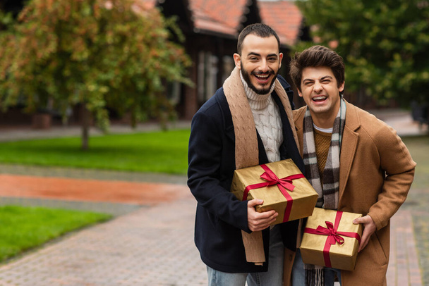 μοντέρνα και χαρούμενα γκέι άνδρες κρατώντας κουτιά δώρων και γελώντας με την κάμερα σε θολή δρόμο - Φωτογραφία, εικόνα
