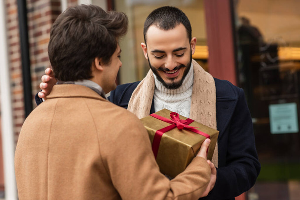 young man holding Christmas present near happy bearded gay partner on city street - Fotó, kép
