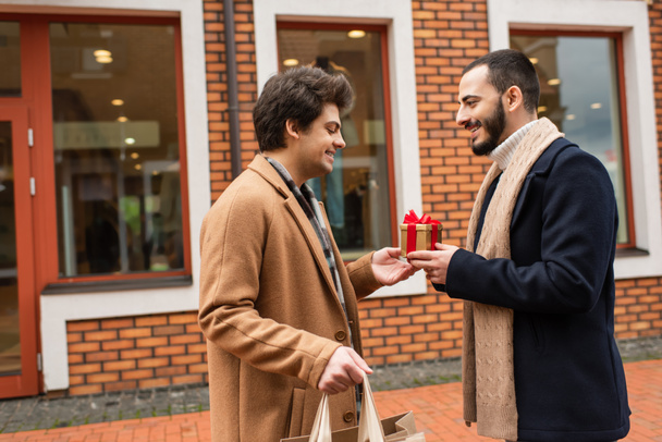 Seitenansicht eines jungen schwulen Mannes mit Einkaufstüten, der einem bärtigen Freund in der Nähe eines Geschäfts auf der Straße ein Weihnachtsgeschenk macht - Foto, Bild