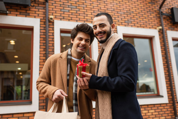 veselý a stylový gayové pár držení nákupní taška a vánoční dárek v blízkosti rozmazané obchod na městské ulici - Fotografie, Obrázek