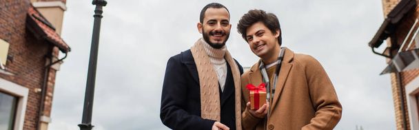 jóvenes y felices hombres gay mirando a la cámara cerca de la pequeña caja de regalo en la calle de la ciudad, pancarta - Foto, imagen