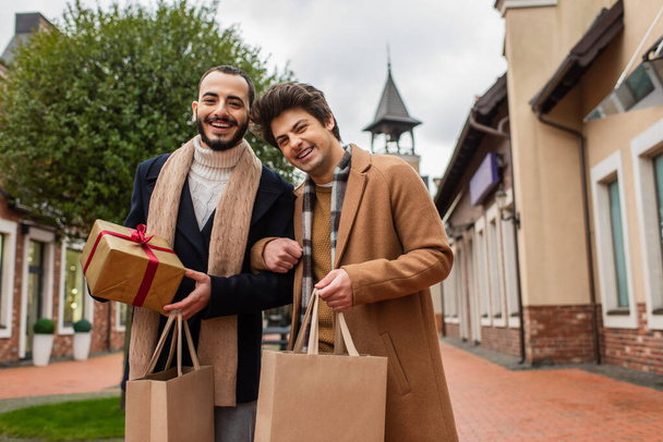 Moda kıyafetleri içinde genç eşcinsel partnerler ellerinde alışveriş çantaları ve Noel hediyeleriyle dışarıda kameralara gülümserken. - Fotoğraf, Görsel