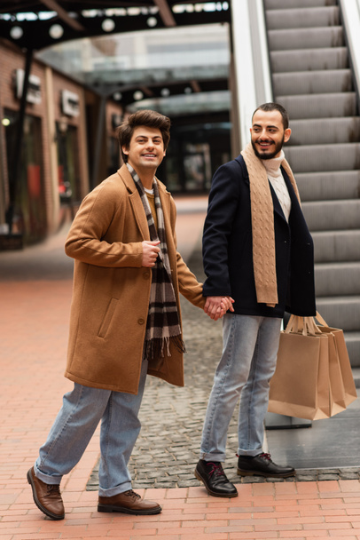 γκέι άνδρας σε μπεζ παλτό και καρό μαντήλι κρατώντας το χέρι με χαμογελαστό γενειοφόρο φίλο με τσάντες για ψώνια κοντά σε κυλιόμενες σκάλες - Φωτογραφία, εικόνα