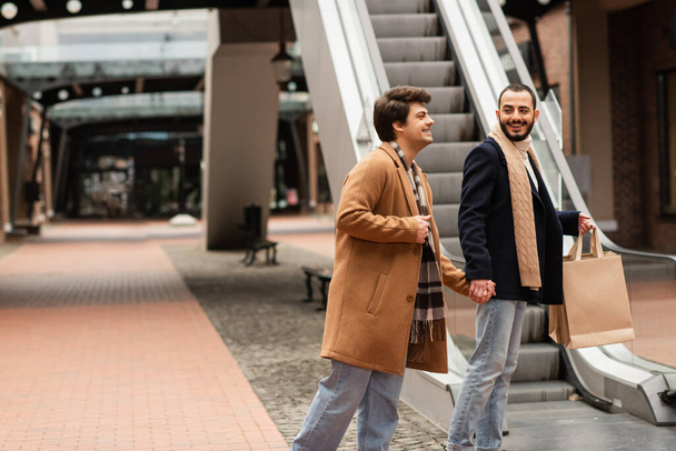 cheerful gay couple in fashionable outfit holding hands while standing with shopping bags near escalator - Φωτογραφία, εικόνα