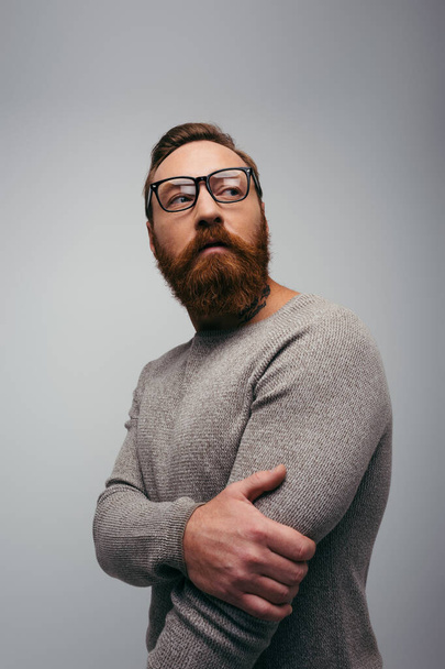 Stílusos szakállas férfi portréja pulóverben és szemüvegben, amint elfordul a szürkén.  - Fotó, kép