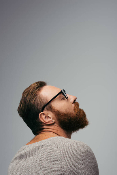 Side view of trendy bearded man in eyeglasses closing eye isolated on grey  - Photo, Image
