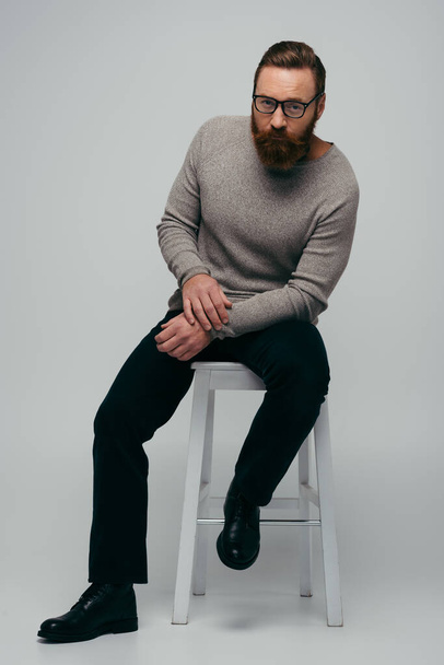 Full length of stylish bearded man in eyeglasses sitting on chair on grey background  - Photo, Image