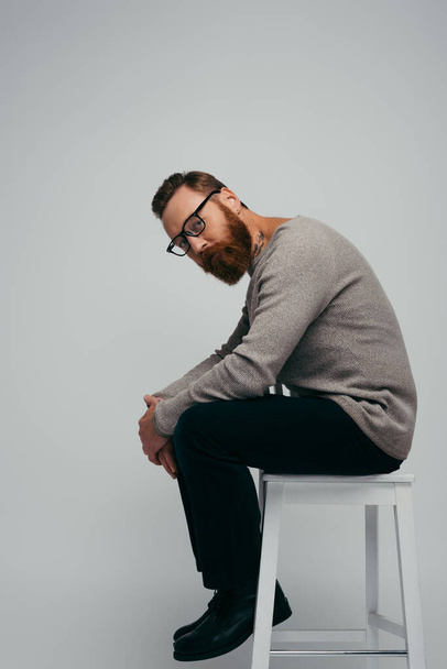 Stylish man in jumper and eyeglasses sitting on chair isolated on grey  - Foto, Bild