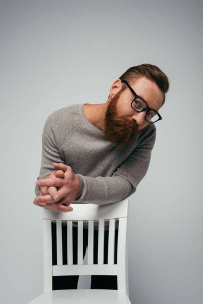 Μοντέρνος άντρας με γυαλιά που κοιτάζει την κάμερα κοντά σε καρέκλα που είναι απομονωμένη σε γκρι  - Φωτογραφία, εικόνα