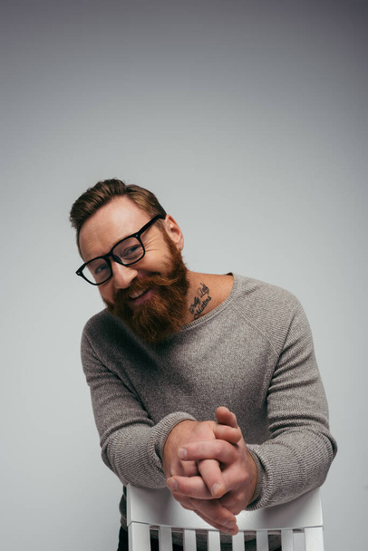 Positive bearded man in eyeglasses looking at camera near chair isolated on grey  - Фото, зображення