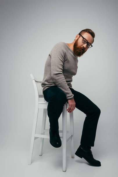 Full length of trendy bearded model in eyeglasses posing on chair on grey background  - Zdjęcie, obraz