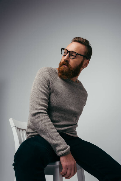 Hombre barbudo de moda en jersey posando en silla sobre fondo gris  - Foto, imagen
