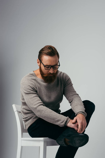 Trendy bearded man in eyeglasses touching leg while sitting on chair on grey background  - Foto, imagen