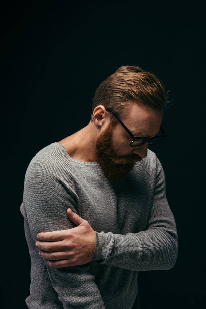 Stylish upset man in eyeglasses touching arm isolated on black  - Fotó, kép