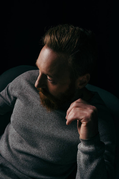 Bearded man in jumper looking away while sitting on armchair isolated on black  - Foto, immagini