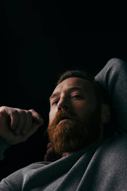Низький кут зору стильного бородатого чоловіка в джемпері, який дивиться на камеру ізольовано на чорному з тіні
  - Фото, зображення