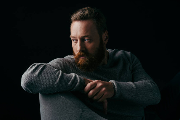 Bearded model in grey jumper looking away while sitting on armchair isolated on black  - Photo, image
