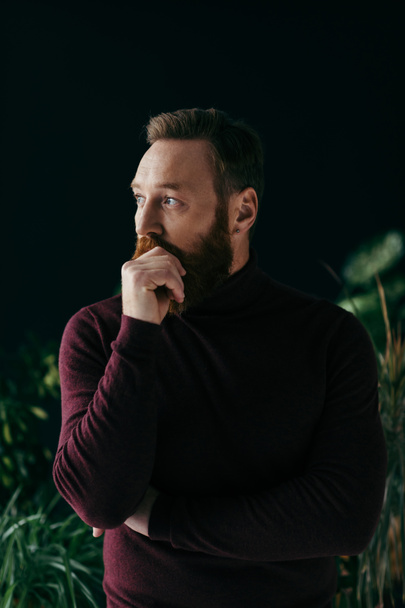 Stylish man in burgundy sweater touching moustache near plants isolated on black  - Foto, imagen