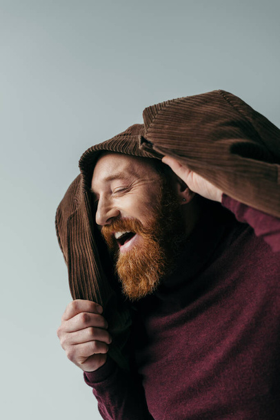 man met baard met bruine blazer op hoofd glimlachend geïsoleerd op grijs  - Foto, afbeelding
