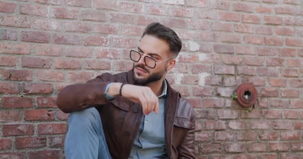 cool guy with stylish hair holding knee up and elbow above, looking to side and adjusting glasses, smiling and sitting outdoor in a medieval town from Romania - Footage, Video