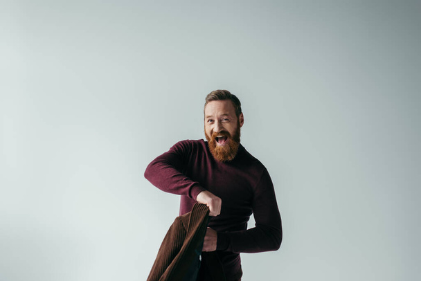 amazed and bearded man in turtleneck holding blazer while looking at camera isolated on grey  - Foto, afbeelding
