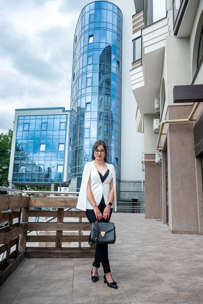 een zakenvrouw in een business style pak staat met een tas in haar handen tegen de achtergrond van een modern kantoorcentrum. Het concept van een zakenvrouw midden in de stad - Foto, afbeelding