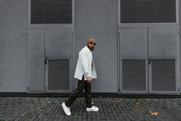 full length of stylish man in beanie hat and sunglasses walking in street  - Foto, afbeelding