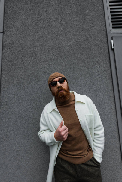 stylish man in beanie hat and sunglasses posing with hand in pocket outside - Foto, Imagen