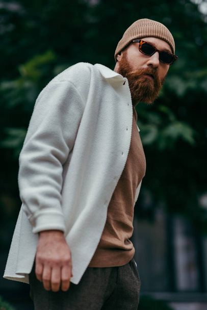 bearded man in stylish sunglasses and beanie hat standing in white shirt jacket near green leaves - Fotó, kép