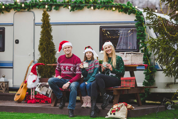 Glückliche kaukasische Familie trinkt Tee oder Kakao vor dem weihnachtlich geschmückten Wohnwagen - Foto, Bild