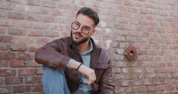 handsome young man with beard holding elbow on knee, fixing eyeglasses and jacket and smiling while looking to side outdoor in an old town - Footage, Video