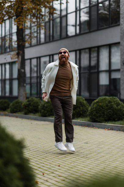 full length of bearded man in beanie hat and trendy sunglasses jumping while smiling near modern building  - Foto, imagen