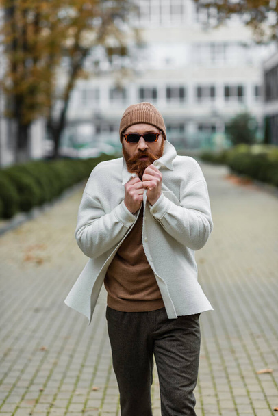 bearded man in beanie hat and trendy sunglasses feeling cold while walking outside - Фото, изображение