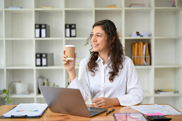 Профессиональная и расслабленная кавказская предпринимательница, размышляющая, глядя в окно, думающая о своем успехе, мечтающая, потягивая кофе в своем офисе. - Фото, изображение