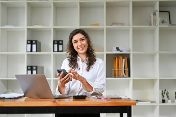 Cheerful millennial caucasian businesswoman or female account executive sitting at her desk, using her phone, smiling and looking at the camera. - Photo, Image