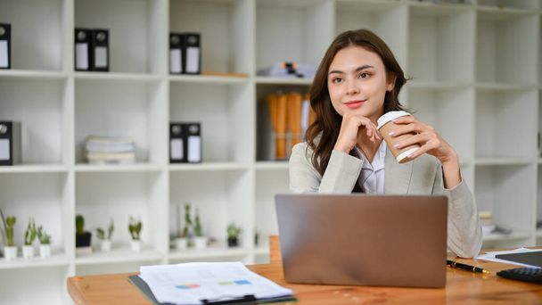 Attractive and charming young caucasian businesswoman in business suit sipping her morning coffee, looking out the window, sitting at her desk. - Photo, Image
