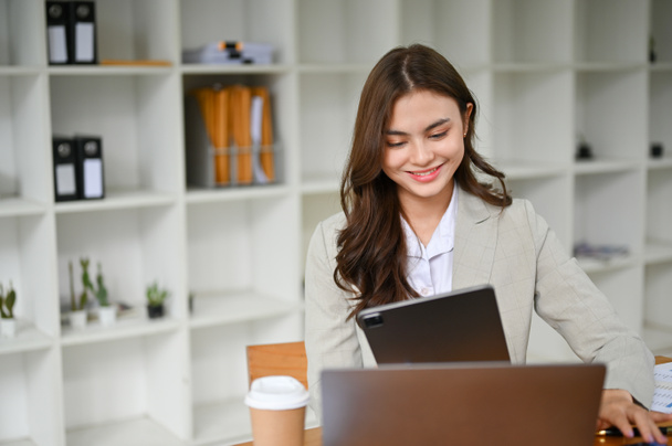 Привлекательная и жизнерадостная молодая кавказская деловая женщина с цифровым планшетом, работающая за своим рабочим столом в современной офисной комнате. - Фото, изображение