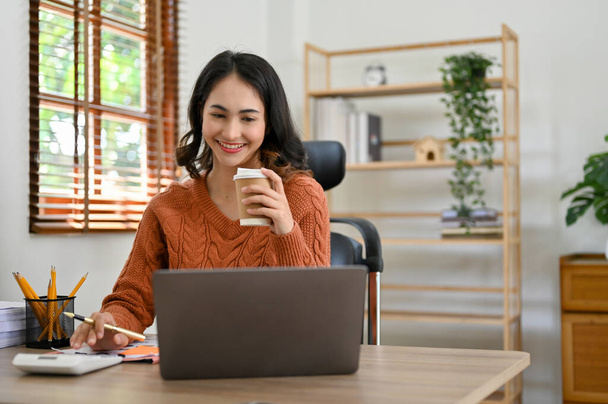 Successful and attractive millennial Asian businesswoman or female manager in casual clothes sipping her morning coffee while working on her tasks on laptop computer in office. - Photo, Image
