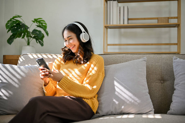 美しい若いアジアの女性は彼女の家のリビングルームでソファでリラックスし、彼女の携帯電話を使用して、ヘッドフォンを介して音楽を聴く. - 写真・画像