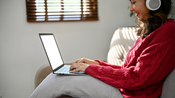 Beautiful young Asian female in red sweater wearing headphones, using laptop to manage her tasks or research online information while sitting on a couch in her living room. rear view - Photo, Image