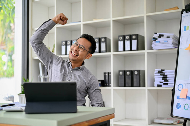Nadšený dospělý asijský podnikatel nebo muž šéf u svého stolu, ukazuje vítěznou ruku, usmívá se, slaví svůj úspěch nebo obchodní soutěž vyhrál. - Fotografie, Obrázek